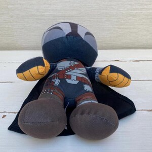 May include: A plush doll depicting a character in a dark blue and gray helmet and armor, with a black cape. The doll has brown legs and yellow and black accents on the arms. The doll is approximately 6 inches tall.