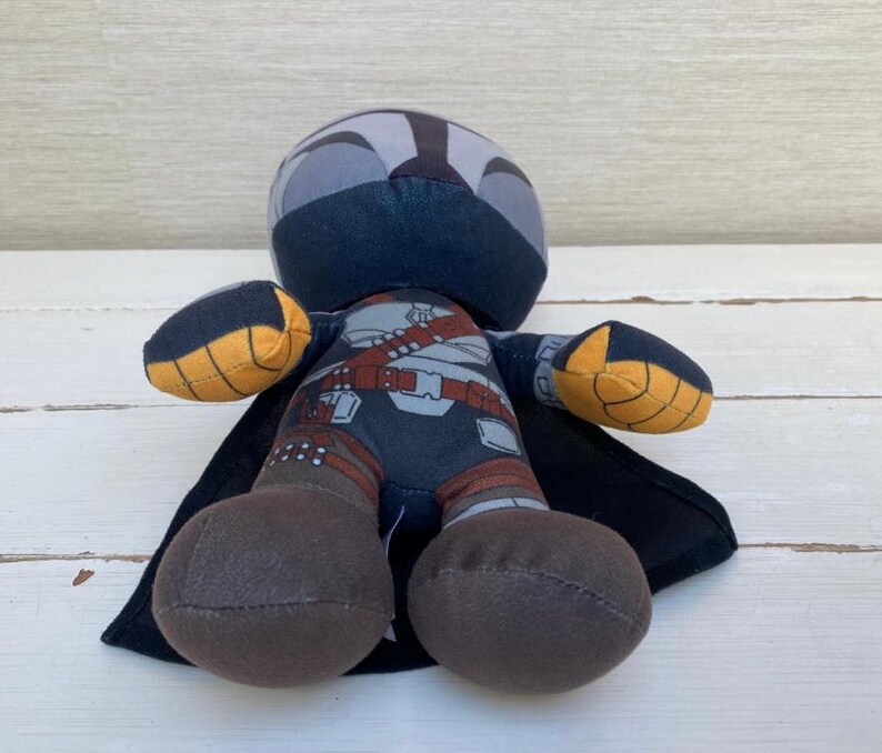 May include: A plush doll depicting a character in a dark blue and gray helmet and armor, with a black cape. The doll has brown legs and yellow and black accents on the arms. The doll is approximately 6 inches tall.
