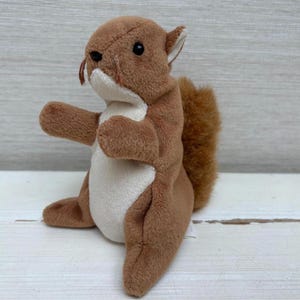 May include: A plush squirrel toy in shades of brown and white. The stuffed animal has a fluffy tail, small black eyes, and a white belly. The squirrel is sitting upright with its arms outstretched.