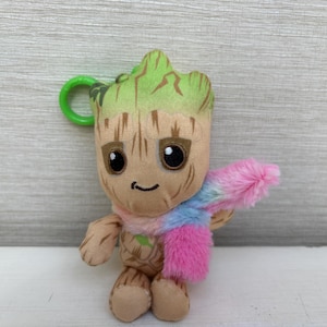 May include: A plush toy keyring featuring a tan, tree-like character with a light green top, large black eyes, and a small smile. The character wears a soft, fuzzy scarf with a multicoloured pattern of pink, blue, and white, and has a green plastic ring.