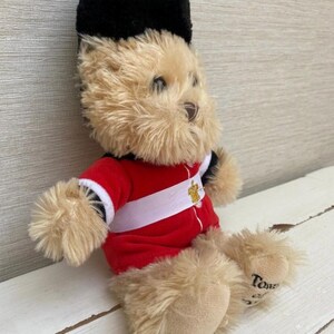 May include: A plush teddy bear dressed as a British Royal Guard. The bear has tan fur, a black hat, and a red jacket with a white stripe. The bear is sitting on a white wooden surface.