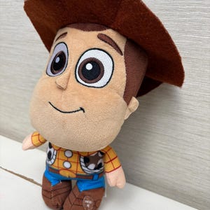May include: A plush toy of the character Woody from Toy Story. The toy has a brown cowboy hat, a tan face, and a yellow shirt with a cow print vest. Blue jeans and brown boots complete the outfit. The toy has the words "PRESS ME" on the boot.