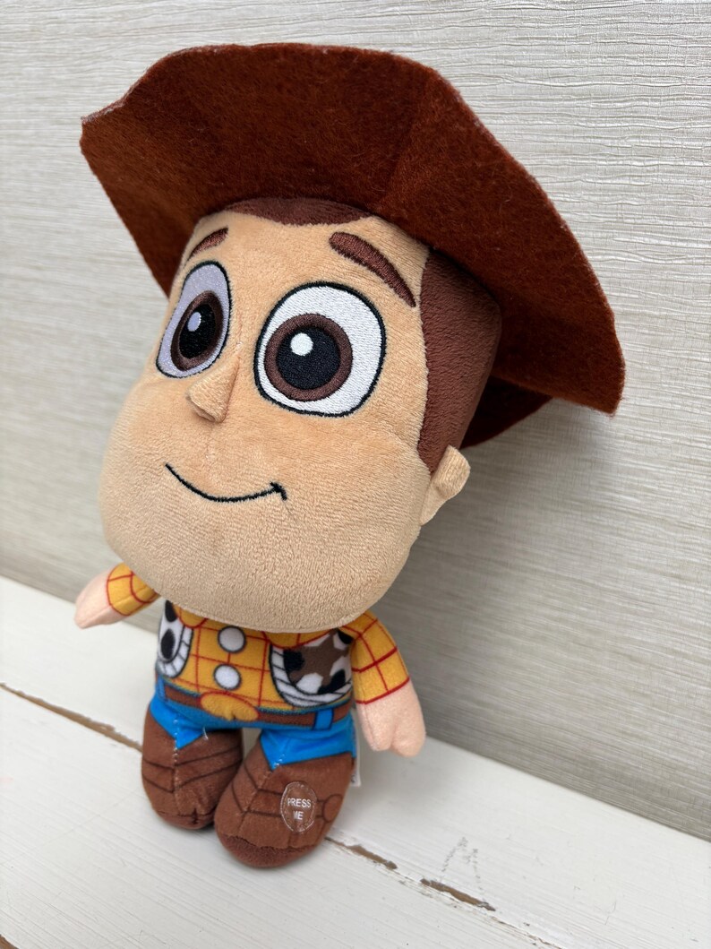 May include: A plush toy of the character Woody from Toy Story. The toy has a brown cowboy hat, a tan face, and a yellow shirt with a cow print vest. Blue jeans and brown boots complete the outfit. The toy has the words "PRESS ME" on the boot.