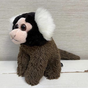 May include: A plush marmoset monkey toy with brown fur, a black head, and white ear tufts. The monkey has large, round eyes and a pink face. The toy is sitting on a white surface.