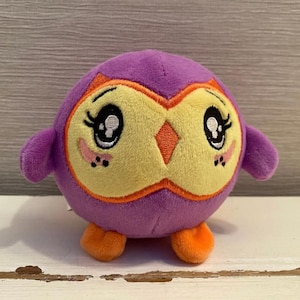 Può includere: Un giocattolo a forma di gufo di peluche, di colore viola, con viso giallo e dettagli arancioni. Il gufo ha grandi occhi, piccole ali e piedi arancioni. Il giocattolo è progettato per essere morbido e coccoloso.