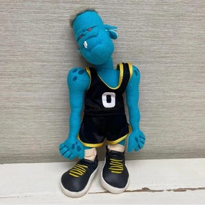 May include: A turquoise monster doll wearing a black basketball vest with the number 0 in white, black shorts, and black trainers with yellow detailing. The doll has red eyes and a single tooth.