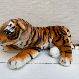 Può includere: Un giocattolo di peluche a forma di tigre in posizione sdraiata. Il giocattolo presenta una pelliccia arancione con strisce nere, zampe bianche e un viso dettagliato con segni neri e bianchi. Il giocattolo è realizzato in materiale morbido.