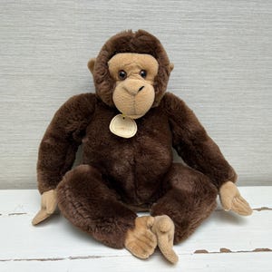 Puede incluir: Un juguete de peluche de mono marrón con cara, manos y pies de color beige. El peluche está sentado con las piernas separadas y los brazos extendidos. Una pequeña etiqueta cuelga de su cuello.