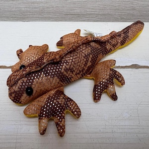 Könnte beinhalten: Ein braunes Stofftier in Form einer Eidechse mit einem gesprenkelten Muster. Das Spielzeug hat eine gelbe Unterseite und schwarze Augen. Die Eidechse liegt auf einer weißen Holzoberfläche.