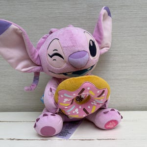 Puede incluir: Un peluche de color lavanda con grandes orejas y una expresi&oacute;n gui&ntilde;adora, sosteniendo una rosquilla amarilla y rosa. El peluche tiene una cara cosida y est&aacute; sentado sobre una superficie blanca.