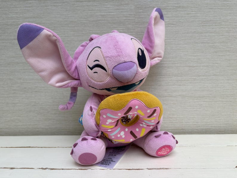 Puede incluir: Un peluche de color lavanda con grandes orejas y una expresi&oacute;n gui&ntilde;adora, sosteniendo una rosquilla amarilla y rosa. El peluche tiene una cara cosida y est&aacute; sentado sobre una superficie blanca.