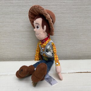 May include: A plush toy of the character Woody from the movie Toy Story. Woody is wearing a brown cowboy hat, a yellow plaid shirt, blue jeans, and brown boots. He has a red neckerchief and a sheriff's badge.