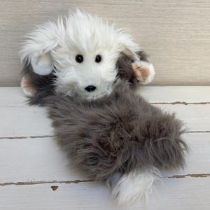 May include: A plush stuffed animal shaped like a dog with long, shaggy fur. The head and tail are white, while the body is a dark grey colour. It features two dark brown eyes, a small black nose, and soft, floppy ears that frame the face.