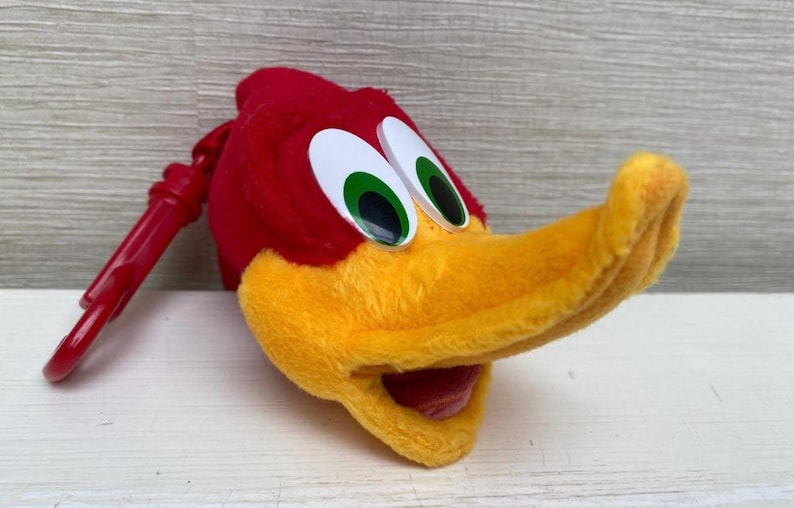 May include: A plush Woody Woodpecker head with a red cap, yellow beak, and large white eyes with green irises. A red plastic clip is attached to the head. The cartoon character is a popular collectible.