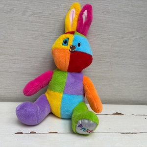 Pode incluir: Um brinquedo de pelúcia em forma de coelho com um design de retalhos multicolorido com segmentos em amarelo, rosa, azul, laranja, vermelho, verde e roxo. O coelho tem orelhas compridas, rosto costurado e a palavra PATCH no pé direito.