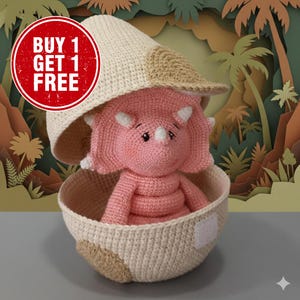 May include: A crocheted pink triceratops dinosaur toy is nestled in a cream and tan crocheted eggshell. The dinosaur has white horns and a frill. The image includes a red circle with the text "BUY 1 GET 1 FREE".