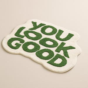 May include: A cream-coloured bath mat featuring the phrase "YOU LOOK GOOD" in green, textured lettering. The mat has an organic, irregular shape. The text is centrally positioned on the soft, fluffy surface.
