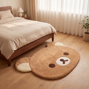 May include: A brown bear-shaped rug with a white and brown face, and two beige ears. The rug is made of a soft, plush material and is placed on a hardwood floor in a bedroom setting.