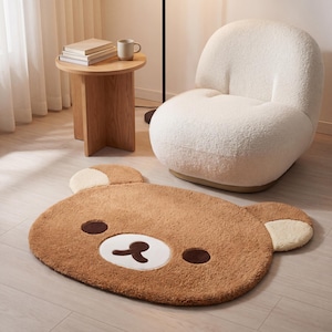 May include: A brown bear-shaped rug with a white mouth and cream-colored ears sits on a light wood floor. A white, fluffy armchair and a small wooden side table with books and a mug are in the background.