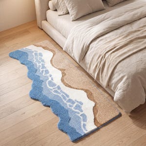 Könnte beinhalten: Ein rechteckiger Teppich mit einem Strandmotiv. Der Teppich zeigt einen blauen Ozean, weiße Wellen und einen sandigen Strand in Beigetönen. Der Teppich liegt auf einem Holzboden neben einem Bett mit beiger Bettwäsche.