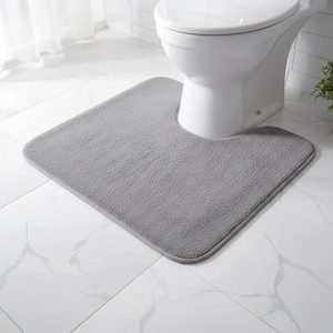 Puede incluir: Una alfombra de baño gris en forma de U con una superficie suave y texturizada. La alfombra está colocada frente a un inodoro blanco sobre un suelo embaldosado. La forma de la alfombra está diseñada para encajar alrededor de la base del inodoro.