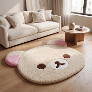 May include: A plush, cream-colored rug shaped like a bear's face, with brown eyes and a white and brown nose. The bear's ears are accented with pink. The rug is on a hardwood floor, near a cream-colored sofa.