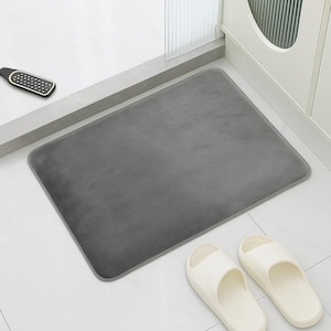 Puede incluir: Una alfombra de baño rectangular gris con esquinas redondeadas se encuentra sobre un suelo de baldosas blancas. Dos sandalias blancas están al lado de la alfombra. También se ve un raspador de pies negro y plateado.