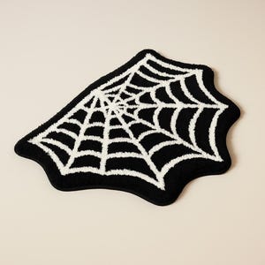 Pode incluir: Um tapete em forma de teia de aranha em preto e branco. O tapete apresenta um fundo preto com um design de teia de aranha branca. Um artigo de decoração para casa com tema de Halloween.