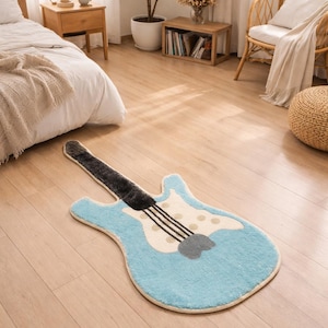 Puede incluir: Una alfombra azul claro con forma de guitarra, con mástil y cuerdas negras. La alfombra tiene un cuerpo color crema con lunares marrones claros y un golpeador gris. Está colocada sobre un suelo de madera clara en un dormitorio.
