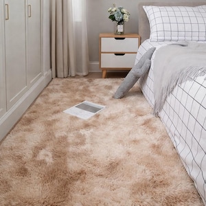 Puede incluir: Una alfombra de pelo largo beige cubre el suelo de un dormitorio. La alfombra tiene una textura suave y esponjosa. Se ve una cama con una colcha y almohada a cuadros, junto con una mesita de noche y un armario. Una revista está sobre la alfombra.