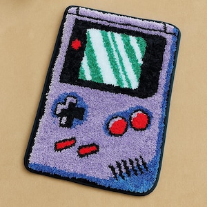 May include: A purple, black, and green Game Boy-shaped rug. The rug features a screen with green and white stripes, a directional pad, red buttons, and a black border. The rug is made of a soft, textured material.