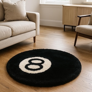 May include: A round black rug with a white number eight design. The rug is placed on a hardwood floor in a living room setting, near a beige sofa and a wooden cabinet.