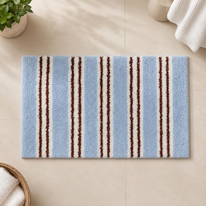 May include: A rectangular bath mat featuring a repeating pattern of vertical stripes in light blue, white, and dark brown. The mat has a soft, tufted texture and is oriented horizontally, displaying a consistent striped design across its surface.