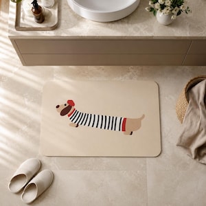 Könnte beinhalten: Eine rechteckige, beige Badematte mit einer stilisierten Illustration eines Dackels mit langem Körper. Der Hund trägt ein rotes Barett und ein schwarz-weiß gestreiftes Shirt. Die Matte hat abgerundete Ecken und eine glatte Oberfläche.