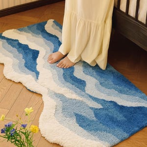 Könnte beinhalten: Ein blau-weißes Wellenmuster-Teppich. Der Teppich hat ein weiches, strukturiertes Aussehen, wobei Blautöne und Weiß einen Farbverlauf erzeugen. Der Teppich liegt auf einem Holzboden.