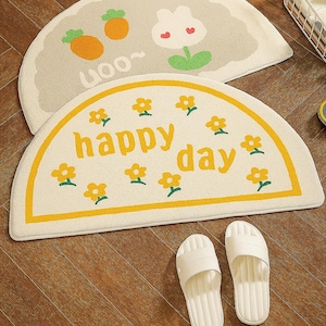 Può includere: Due tappeti a semicerchio con disegni di cartoni animati e un paio di sandali bianchi. Un tappeto ha carote e un motivo floreale, l'altro dice "happy day" con fiori gialli. I sandali sono su un pavimento in legno.