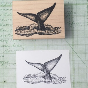 May include: A black and white illustration of a whale tail splashing in the water. The image is printed on a wooden block.