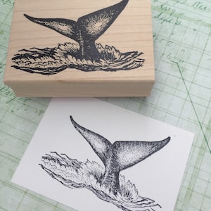 May include: A black and white illustration of a whale tail splashing in the water. The image is printed on a wooden block.
