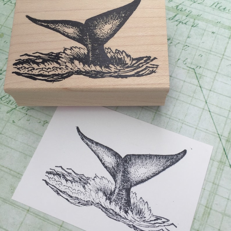May include: A black and white illustration of a whale tail splashing in the water. The image is printed on a wooden block.