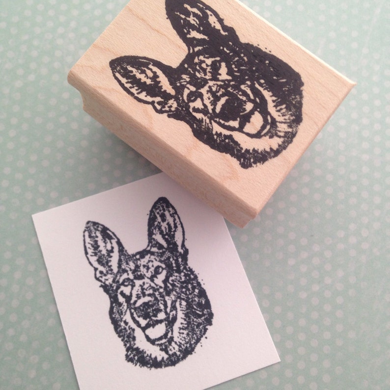 Puede incluir: Un sello de goma de madera con una ilustraci&oacute;n en blanco y negro de la cabeza de un perro pastor alem&aacute;n. El perro mira hacia la derecha y tiene la boca abierta.