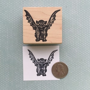 May include: A wooden block stamp with a black and white illustration of a gargoyle with wings. The gargoyle is facing forward with its wings spread out. The stamp is about the size of a US quarter.