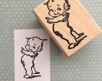 以下が含まれることがあります： 右を指さす翼のある天使の白黒の線画。画像はゴム印である木製のブロックに印刷されています。