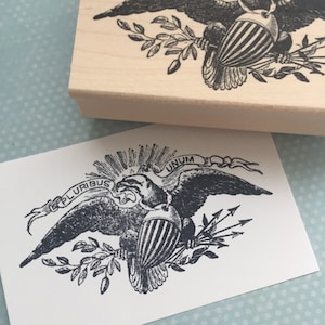 May include: A black and white illustration of an eagle with outstretched wings holding a bundle of arrows and an olive branch. The eagle is perched on a branch and has a banner above it that reads "E PLURIBUS UNUM".