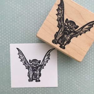 May include: A black and white rubber stamp with a detailed illustration of a gargoyle with wings. The gargoyle is facing forward with its wings spread out. The stamp is on a wooden block.