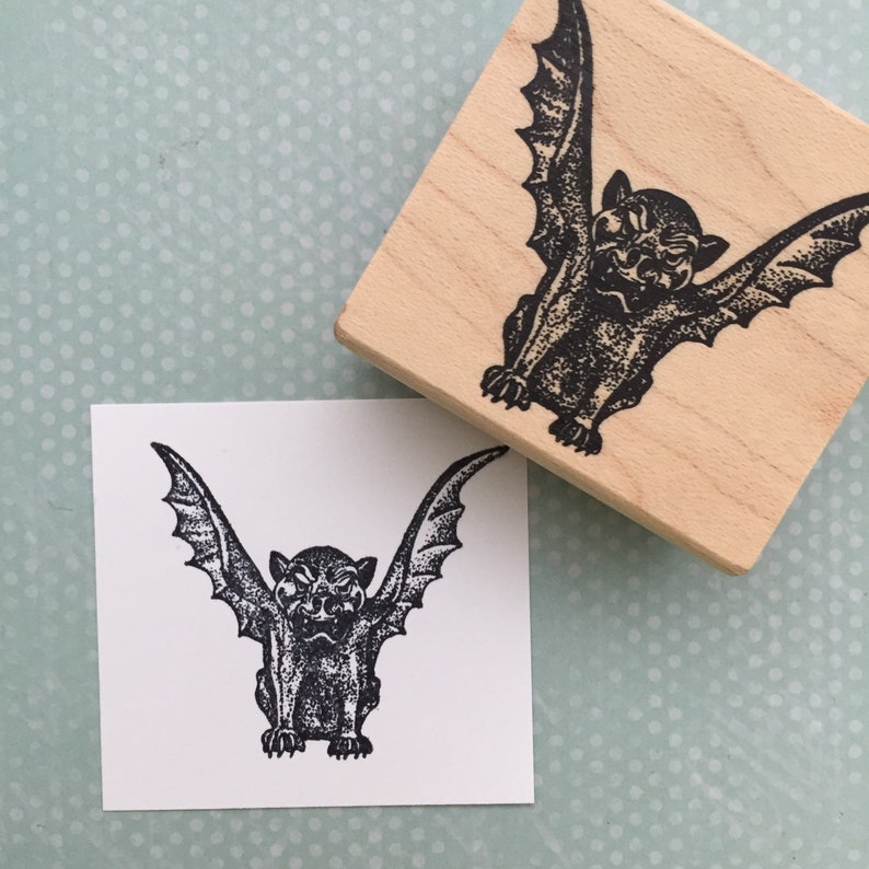 May include: A black and white rubber stamp with a detailed illustration of a gargoyle with wings. The gargoyle is facing forward with its wings spread out. The stamp is on a wooden block.