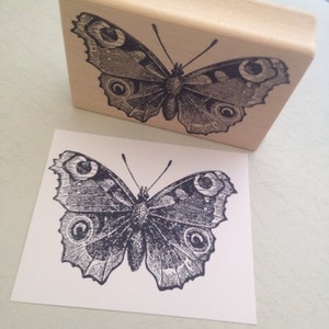 Puede incluir: Un sello de goma negro y blanco de una mariposa con detalles intrincados. La mariposa tiene grandes ocelos redondos en sus alas.