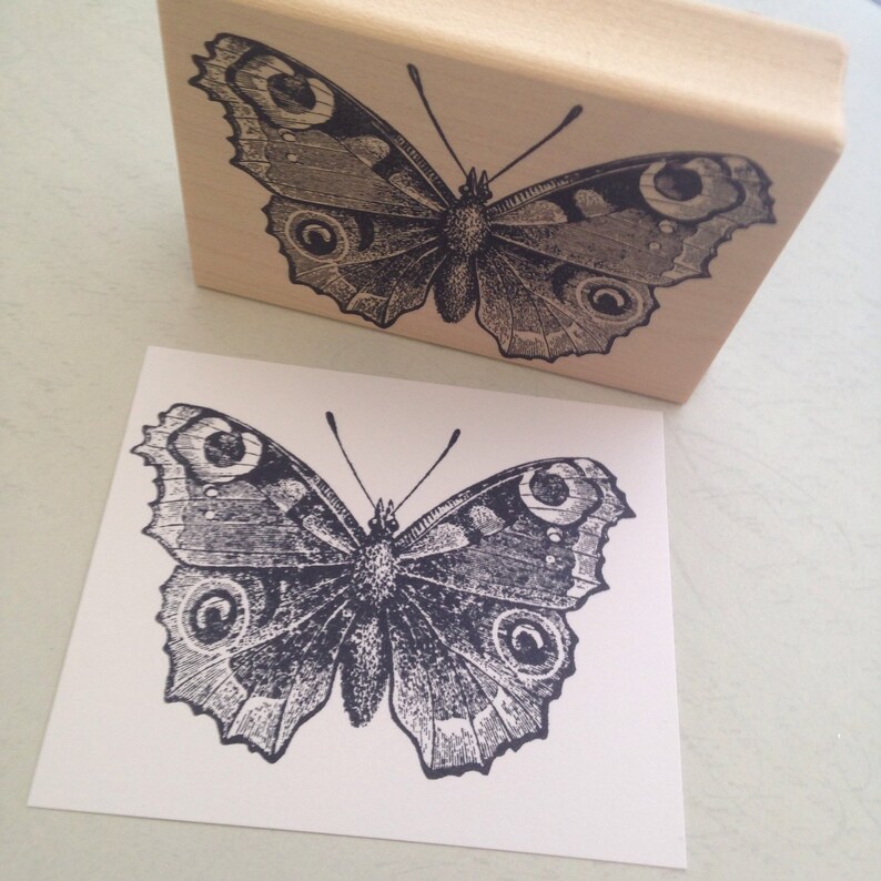Puede incluir: Un sello de goma negro y blanco de una mariposa con detalles intrincados. La mariposa tiene grandes ocelos redondos en sus alas.
