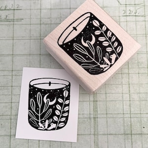 May include: A wooden stamp with a black and white candle design. The candle illustration features a crescent moon, stars, and botanical elements. A printed image of the design sits next to the stamp on a grid background.