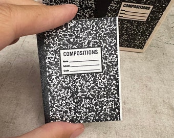 Könnte beinhalten: Ein schwarz-weißes Kompositionsheft mit dem Wort "COMPOSITIONS" auf dem vorderen Umschlag. Das Heft hat Zeilen für Name, Schule und Klasse.