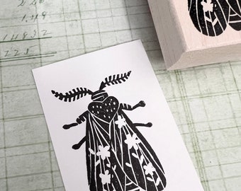 May include: A black and white insect stamp with a floral design. The stamp is on a wooden block. A printed image of the insect is next to the stamp on a white piece of paper. The background is a grid pattern.